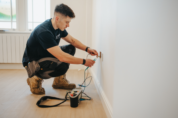 An electrician bent down, testing a plug socket in a wall with a machine.
